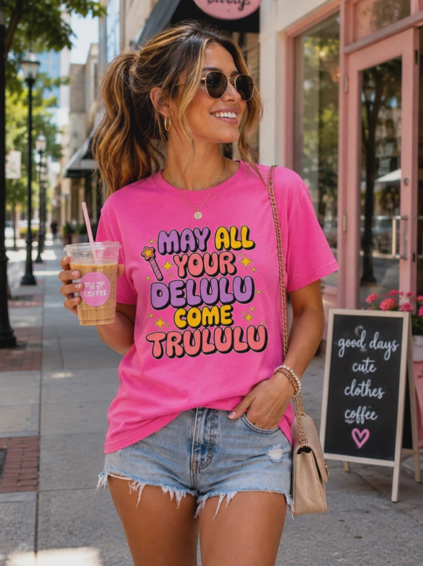 Animated lifestyle video of a woman wearing a bright pink "May All Your Delulu Come Trululu" graphic T-shirt outside a trendy coffee shop. She looks down to admire the colorful retro design, smiles, then looks up at the camera while holding an iced coffee, highlighting the playful unisex graphic tee in a stylish everyday setting.