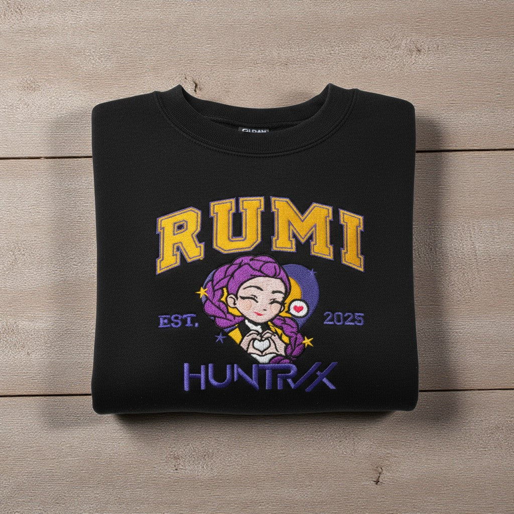 Folded black sweatshirt with 'RUMI HUNTRIX' design on a wooden surface