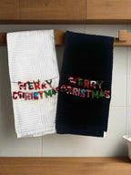 Pair of kitchen towels staged in kichen with the words Merry Christmas with festive colored font.