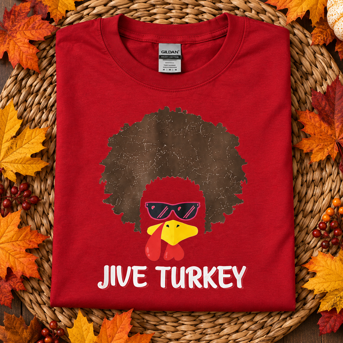 A folded red t-shirt with a 'Jive Turkey' graphic print, displayed on a woven straw mat surrounded by autumn leaves. The graphic features a brown afro, a yellow turkey head with sunglasses, and white text.