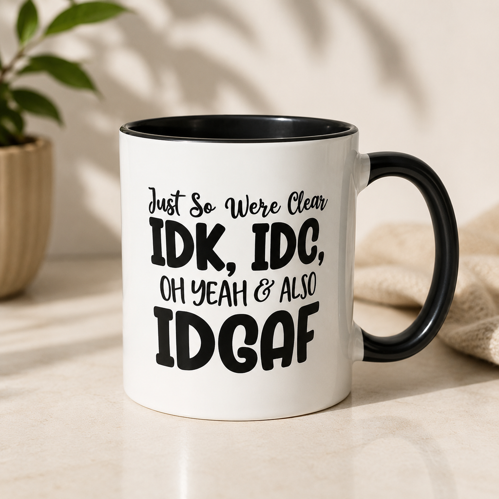 A white ceramic mug with a black interior and black handle, featuring the text 'Just So Were Clear IDK, IDC, OH YEAH & ALSO IDGAF' in black lettering. The mug is set on a light beige surface with a blurred plant and neutral fabric in the background.