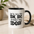 A white ceramic mug with a black interior and black handle, featuring the text 'Just So Were Clear IDK, IDC, OH YEAH & ALSO IDGAF' in black lettering. The mug is set on a light beige surface with a blurred plant and neutral fabric in the background.
