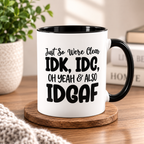 A white ceramic mug with a black handle and black interior, printed with the text 'Just So We Were Clear IDK, IDC, OH YEAH & ALSO IDGAF' in black lettering. The mug sits on a round wooden coaster on a wooden table, with a potted plant and books blurred in the background.