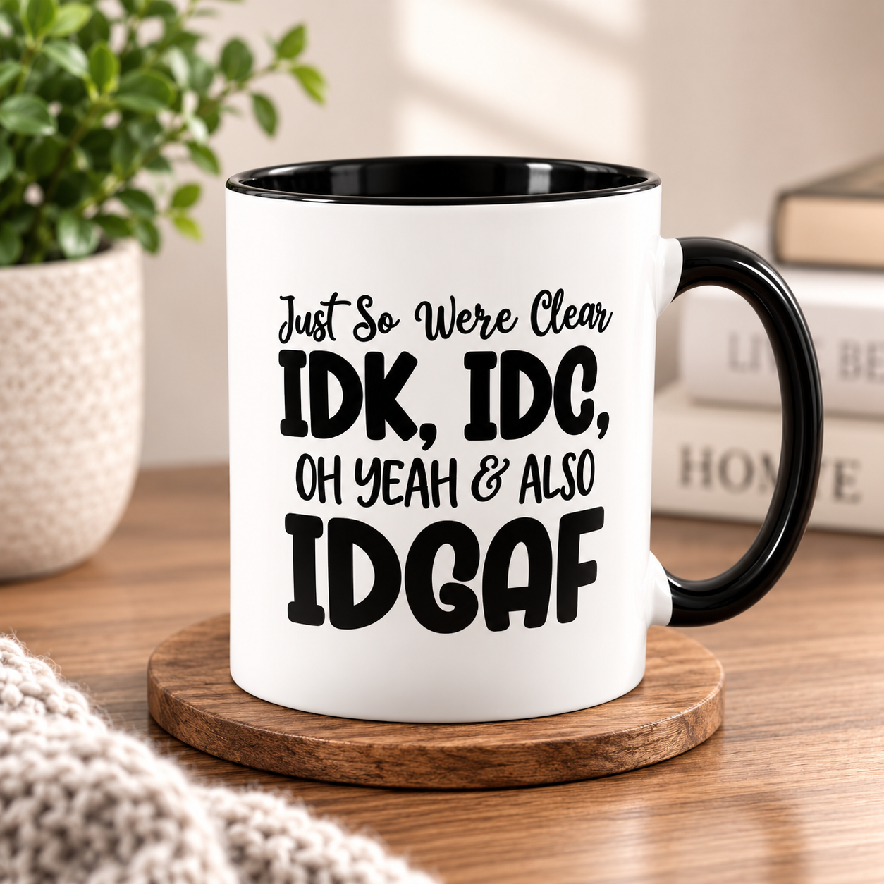 A white ceramic mug with a black handle and black interior, printed with the text 'Just So We Were Clear IDK, IDC, OH YEAH & ALSO IDGAF' in black lettering. The mug sits on a round wooden coaster on a wooden table, with a potted plant and books blurred in the background.