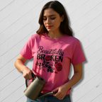 Woman wearing a pink t-shirt with 'Beautifully Broken' design, holding a watering can against a white wooden background.