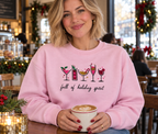 A woman in a cozy cafe setting wearing a light pink crewneck sweatshirt with a holiday-themed cocktail graphic and the text 'full of holiday spirit'. She is holding a white coffee mug with latte art.