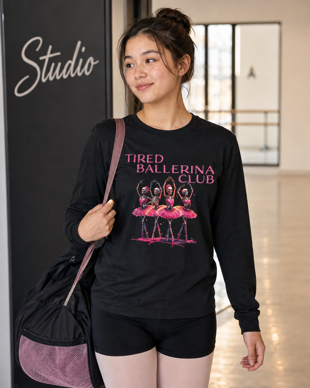 A woman wearing a black long-sleeve graphic t-shirt with pink text reading 'TIRED BALLERINA CLUB' and an illustration of ballerinas, paired with black shorts and carrying a black and pink bag, standing in a studio environment.