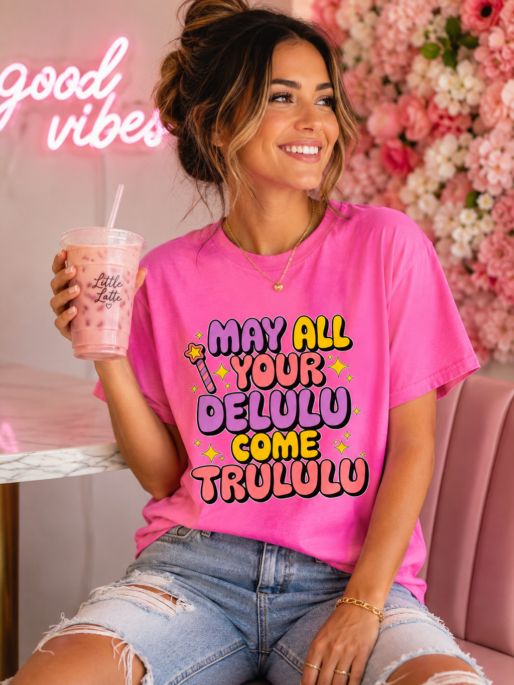 A woman wearing a bright pink graphic t-shirt with the text 'MAY ALL YOUR DELULU COME TRULULU' in colorful bubble letters, holding a pink drink in a pink-themed setting with a floral wall and neon sign.