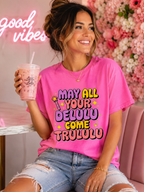 A woman wearing a bright pink graphic t-shirt with the text 'MAY ALL YOUR DELULU COME TRULULU' in colorful bubble letters, holding a pink drink in a pink-themed setting with a floral wall and neon sign.