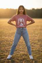 A young woman stands in a grassy field at sunset wearing a dusty rose graphic t-shirt with a white sketch of a woman and the text 'Yall need Jesus', paired with light-wash blue jeans and white sneakers.
